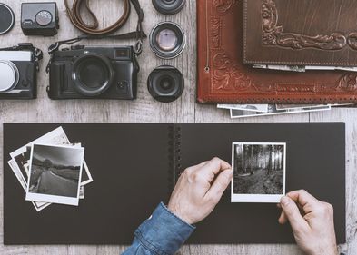 Es liegen verschiedene Kameras und Objektive in einer Ecke. Eine Person hält eine schwarz-weiß Fotografie. Weitere schwarz-weiß Fotografien liegen auf einem leeren Fotoalbum. Alte Leder-Fotoalben liegen in der oberen rechten Ecke.