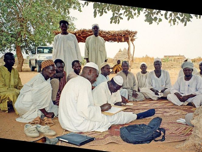 Men sitting in an open circle on carpets. In the background there two men are standing and there are buildings with straw roofs, a white car and a tree.