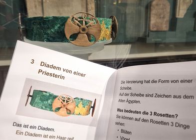 Diadem aus Kupfer im Hintergrund, davor aufgeschlagenes Heft mit Text.