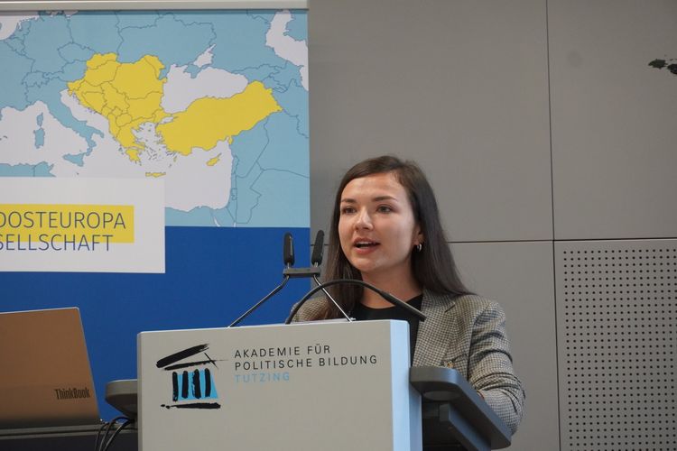 Olga Bostan steht hinter einem Podium. Auf dem Podium steht "Akademie für politische Bildung Tutzing". Im Hintergrund ist eine Landkarte sichtbar, auf der die Region Südosteuropa gelb hervorgehoben ist. Unter der Karte steht "Südosteuropa Gesellschaft".