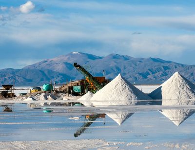 A flat lake is visible in which salt is accumulated to small piles. Behind those piles big machinery is positioned to extract the salt out of the water. A mountain range and the blue sky are visible in the background.